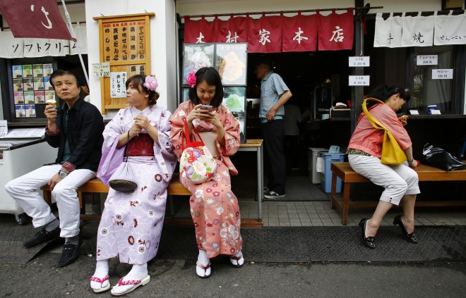 In this photo taken Wednesday, May 6, 2015, foreign tourists wearing traditional Japanese kimonos rest on a bench in front of a Japanese confectionary store near Sensoji Temple in Tokyo. The cheap yen, relaxed visa restrictions and other initiatives are luring travelers from around the globe eager to stretch their budgets and see some UNESCO World Heritage sites, bringing in welcome cash, and myriad complications. (AP Photo/Shizuo Kambayashi)