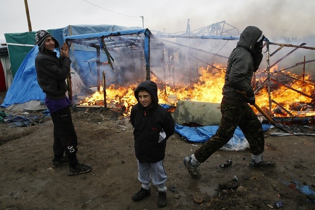 Migrants stand in front of  a burning dwelling in a makeshift  migrants camp near Calais, France,  Tuesday March 1, 2016. The source of the fire is not known.  French authorities have begun dismantling part of the sprawling camp locally referred to as "the jungle" where thousands are hanging out, hoping to make their way to a better life in Britain. (AP Photo/Jerome Delay)