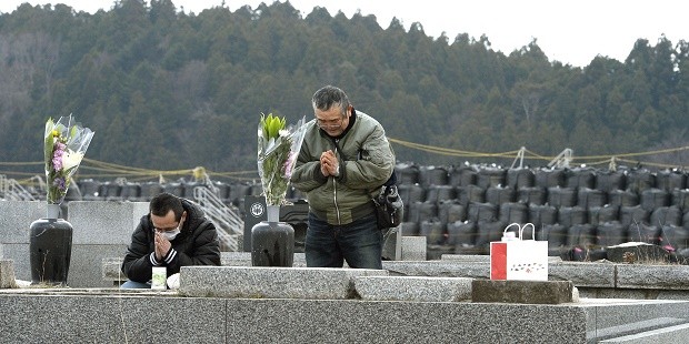 People pray in front of ancestors' grave as black plastic bags containing radiation contaminated waste are piled in the background, in Futaba, Fukushima prefecture, northern Japan Friday, March 11, 2016.  Japan on Friday marked the fifth anniversary of the 2011 tsunami that killed more than 18,000 people and left a devastated coastline along the country's northeast that has still not been fully rebuild. AP