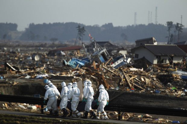 FILE - In this April 15, 2011 file photo, Japanese police officers in protective suits carry a victim at a tsunami-devastated area in the town of Namie as towers of the crippled Fukushima Dai-ichi nuclear power plant are seen in the distance, top right, in Fukushima Prefecture, northeastern Japan. One month after the disaster struck, we slipped into the evacuation zone around the Fukushima Dai-ichi nuclear facility. We approached as near as we dared, just a few kilometers from the plant, and through my viewfinder I saw a scene straight from a science fiction movie: Men in white spacesuits carrying body bags across the gray ash-like wasteland left by the tsunami. Curtains flapping from windows of washed-up bungalows, beached fishing boats and a crumbled road filled the foreground. In the distance were the iron chimneys of the nuclear plant. The scene captured all aspects of the triple disaster, and as I stepped back from my camera I thought, "Wow, Ive got it all in one frame." - Miles Edelsten, former senior video producer.  (AP Photo/Hiro Komae, File)