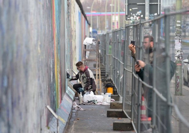 A worker removes graffiti from a painting behind a temporary fence on the East Side Gallery, a much-visited stretch of the Berlin wall, in Berlin on November 5, 2015. The city of Berlin has decided to erect a 80cm high permanent barrier to discourage people from defacing the works made by artists who decorated the yet untouched east side with artwork and political statements after the wall was taken down in 1989-1990. Though designated as a historical landmark, the paintings have yet again been covered in grafitti.   AFP PHOTO / JOHN MACDOUGALL / AFP PHOTO / JOHN MACDOUGALL