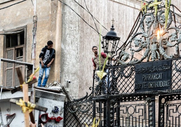Two men stand atop the fence of the Coptic Orthodox Patriarchate in Alexandria as other Egyptians gather outside, after a bomb blast struck outside as worshippers attended Palm Sunday mass on April 9, 2017. The Interior ministry said Coptic Pope Tawadros II was inside the church leading a Palm Sunday service when the suicide bomber was stopped by police outside and blew himself up. A church official said Tawadros had already left the church when the bombing took place. / AFP PHOTO / MOHAMED EL-SHAHED