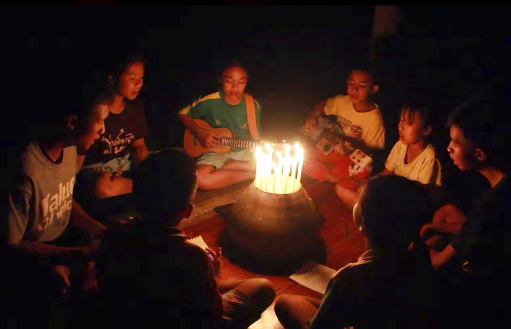 In this image from video made available on Friday, June 29, 2018, a group of young children sit around candles and play a song at a school in Buriram, eastern Thailand, in support for the missing soccer team and their coach in a flooded cave in the north. With songs, prayers and artwork, Thais are keeping hope alive that 12 boys and their soccer coach who disappeared in a flooded mountain cave a week ago will be rescued. (Lek Nai Tung Kwang school via AP)