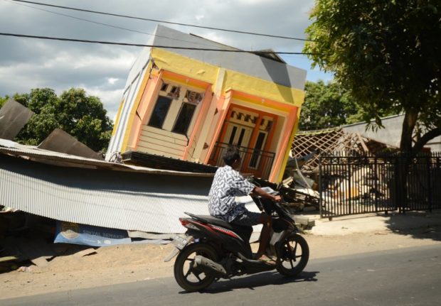 A man riding a motorcycle passes by a damaged house at Sira village in northern Lombok in West Nusa Tenggara province on August 7, 2018, two days after the area was struck by an earthquake. The shallow 6.9-magnitude quake killed at least 98 people and destroyed thousands of buildings in Lombok on August 5, just days after another deadly tremor surged through the holiday island and killed 17. / AFP PHOTO / SONNY TUMBELAKA