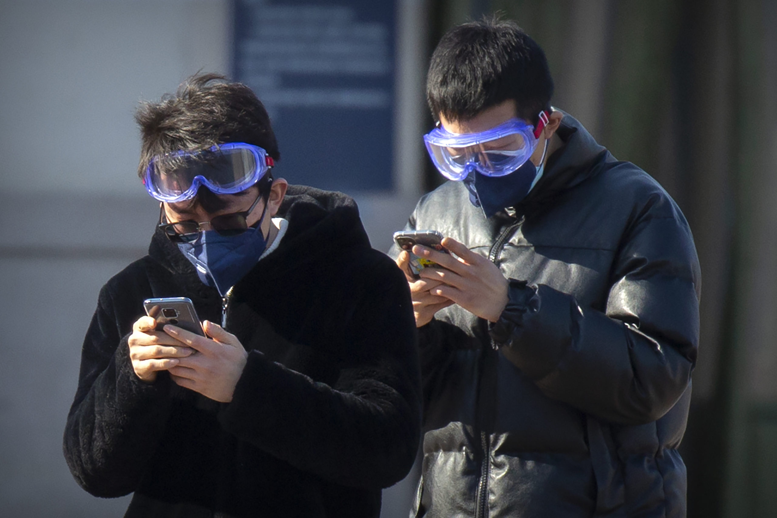 Travelers wear face masks and goggles as they use their smartphones outside the Beijing Railway Station in Beijing, Saturday, Feb. 15, 2020. People returning to Beijing will now have to isolate themselves either at home or in a concentrated area for medical observation, said a notice from the Chinese capital's prevention and control work group published by state media late Friday. (AP Photo/Mark Schiefelbein)