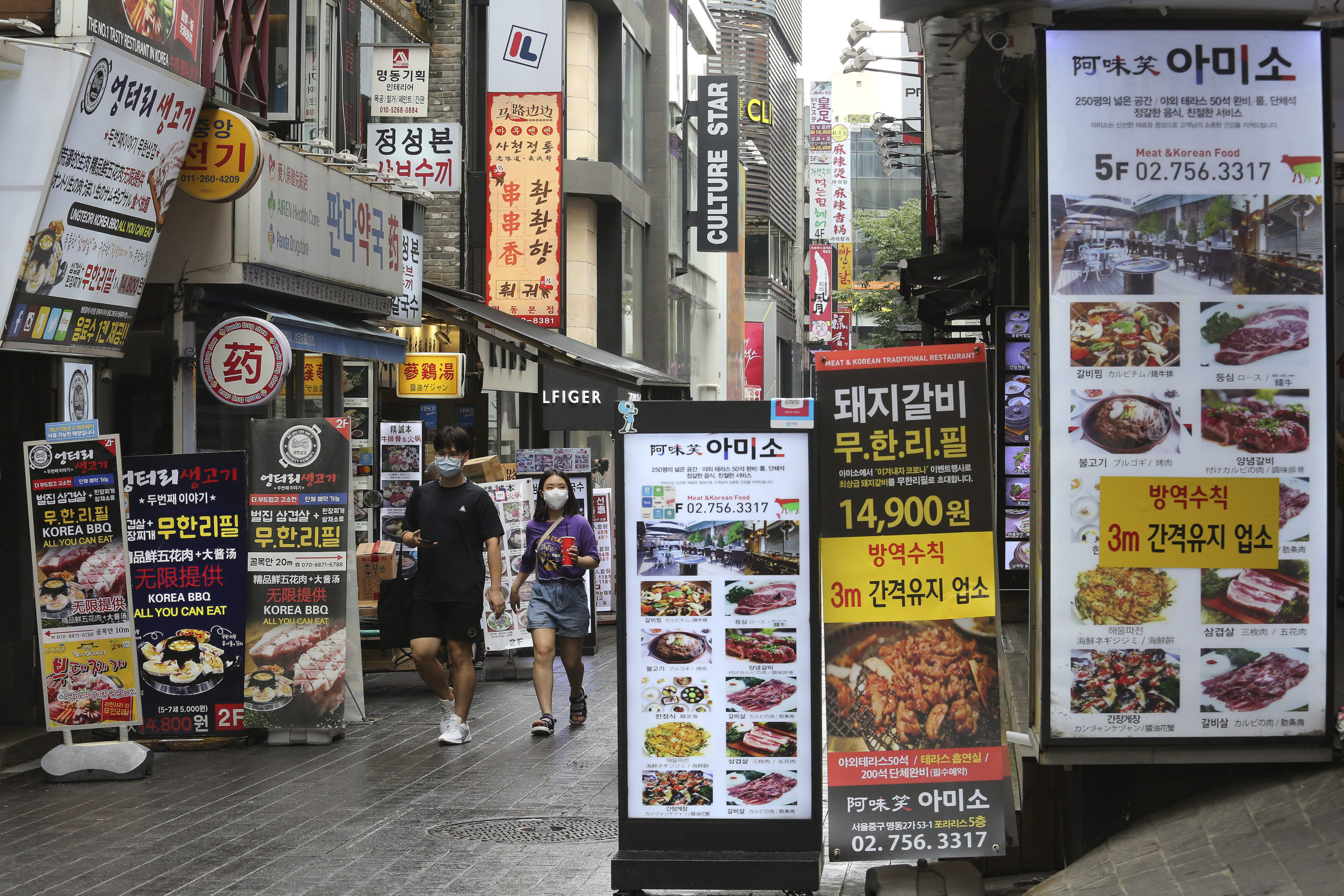 People wearing face masks to help protect against the spread of the coronavirus pass by banners showing the menu items of restaurants in Seoul, South Korea, Friday, Aug. 28, 2020. South Korean officials are considering reducing working hours of restaurants and cafes as the country counted its 15th straight day of triple-digit jumps in coronavirus infections. (AP Photo/Ahn Young-joon)