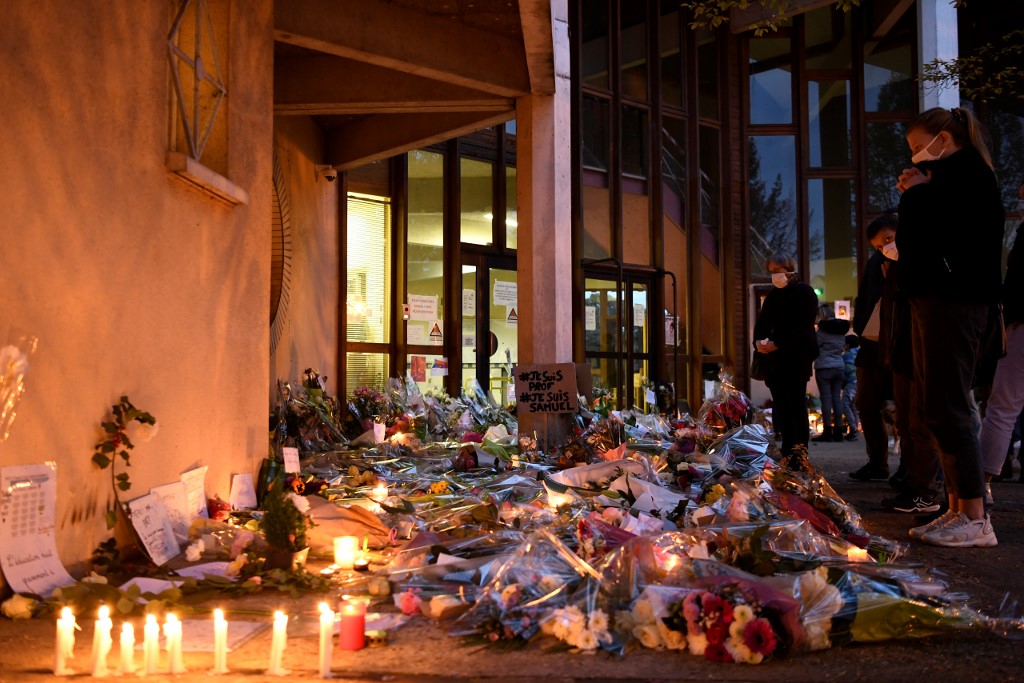 People stand in front of flowers and candles next to a placard reading 'I am a teacher, I am Samuel' at the entrance of a middle school in Conflans-Sainte-Honorine, 30kms northwest of Paris, on October 17, 2020, after a teacher was decapitated by an attacker who has been shot dead by policemen. - The man suspected of beheading on October 16 ,2020 a French teacher who had shown his students cartoons of the prophet Mohammed was an 18-year-old born in Moscow and originating from Russia's southern region of Chechnya, a judicial source said on October 17. Five more people have been detained over the murder on October 16 ,2020 outside Paris, including the parents of a child at the school where the teacher was working, bringing to nine the total number currently under arrest, said the source, who asked not to be named. The attack happened at around 5 pm (1500 GMT) near a school in Conflans-Sainte-Honorine, a western suburb of the French capital. The man who was decapitated was a history teacher who had recently shown caricatures of the Prophet Mohammed in class. (Photo by Bertrand GUAY / AFP)