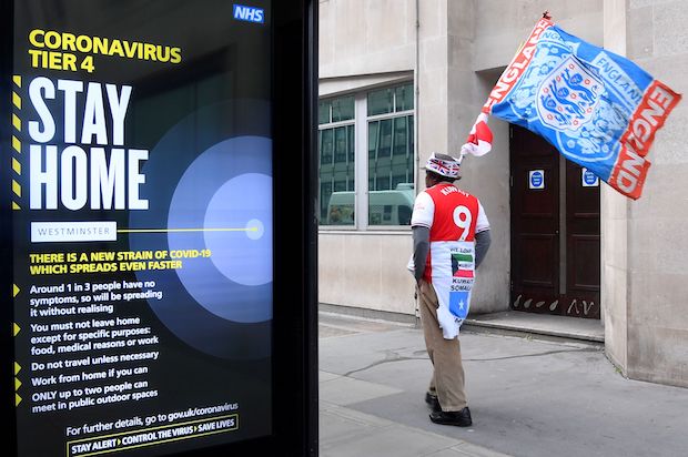 Man with flag walks past government building in London with COVID-19 sign on foreground.