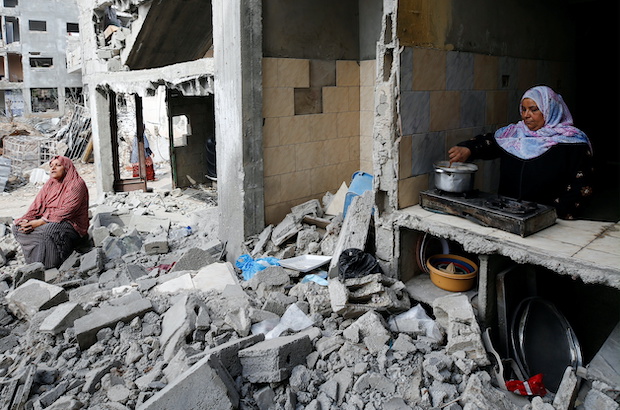A Palestinian woman cooks as another sits amid the rubble of their houses destroyed by Israeli air strikes