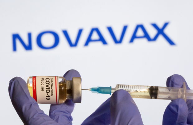 A woman holds a small bottle labeled with a "Coronavirus COVID-19 Vaccine" sticker and a medical syringe in front of displayed Novavax logo in this illustration
