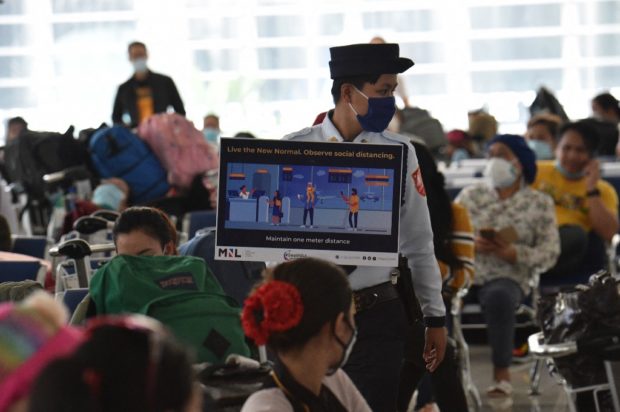 A security guard holding a placard reminding people of social distancing walks past Philippine overseas workers who were quarantined for weeks after returning home as they wait for flights back to their home cities around the country, at Manila's international airport on May 28, 2020. - The Philippine government set up local flights to send thousands of migrant workers stuck in quarantine facilities in Manila back home in an effort to free up crowded quarantine facilities, ahead of new arrivals from overseas. (Photo by Ted ALJIBE / AFP)