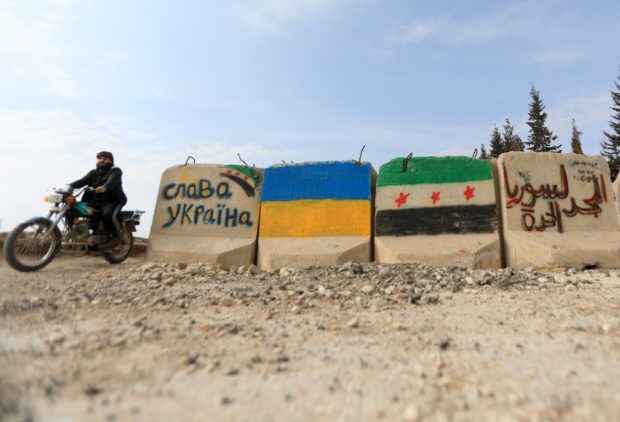 A man rides a motorcycle past cement blocks bearing flags of Ukraine (C L) and the Syrian opposition with inscriptions, one in Cyrillic (L) which reads "Slava Ukraini" ("glory to Ukraine"), a Ukrainian national salute, and another in Arabic which reads "glory to free Syria", near the Syrian rebel-held city of al-Bab in the northern Aleppo governorate, on February 28, 2022. (Photo by Bakr ALKASEM / AFP)