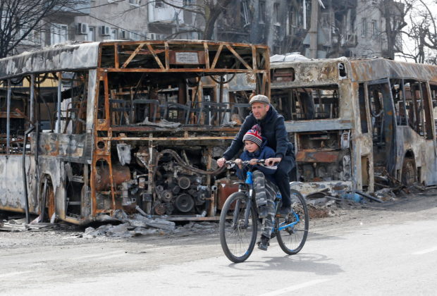 A man and a child ride a bicycle past burnt out buses during Ukraine-Russia conflict in the southern port city of Mariupol, Ukraine April 19, 2022. REUTERS/Alexander Ermochenko