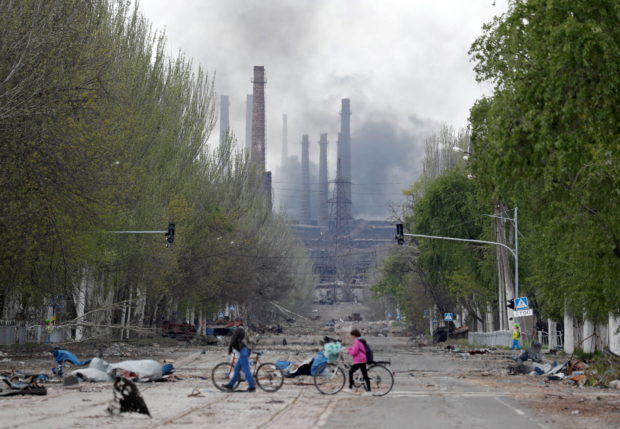 People walk their bikes across the street as smoke rises above a plant of Azovstal Iron and Steel Works during Ukraine-Russia conflict in the southern port city of Mariupol, Ukraine May 2, 2022. REUTERS/Alexander Ermochenko