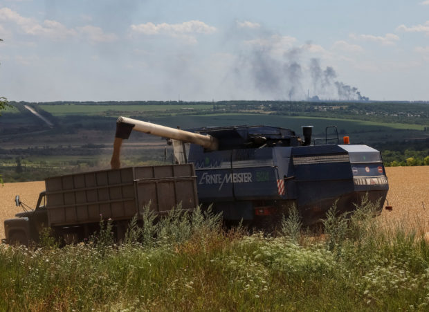 Farmers harvest wheat as Vuhlehirsk's heat power plant burns in the distance after a shelling, amid Russia's attack on Ukraine, in the Donbas region, Ukraine July 13, 2022. REUTERS/Gleb Garanich