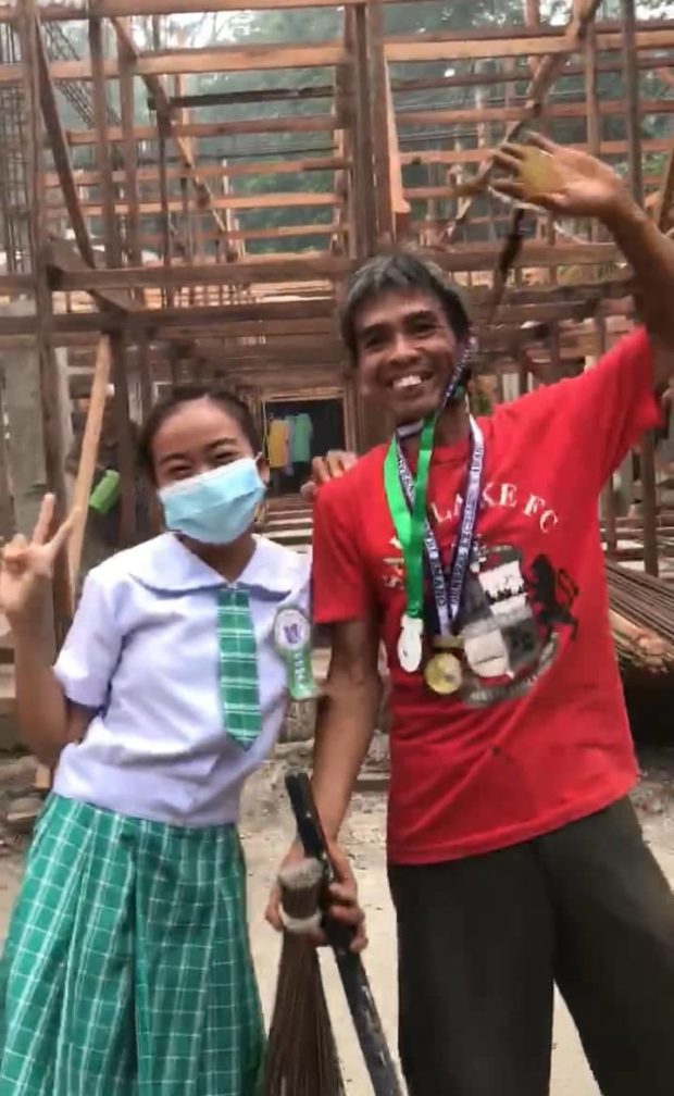 Achiever daughter wearing school uniform posed for a picture beside her construction worker dad wearing red t-shirt and her daughter's medals