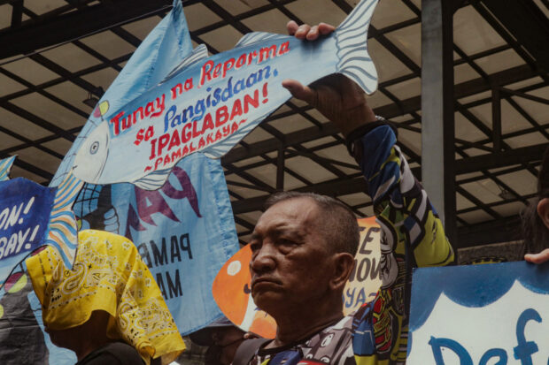 Mang Nic Miane, a fisherman from Cavite and PAMALAKAYA member, holds a placard that reads ‘Tunay na Reporma sa Pangisdaan, ipaglaban!’ at a rally in front of the SMC head office on Saturday. He and his grandchildren came to Mandaluyong on Earth Day to protest the SMC reclamation projects. / Sofia Abrogar