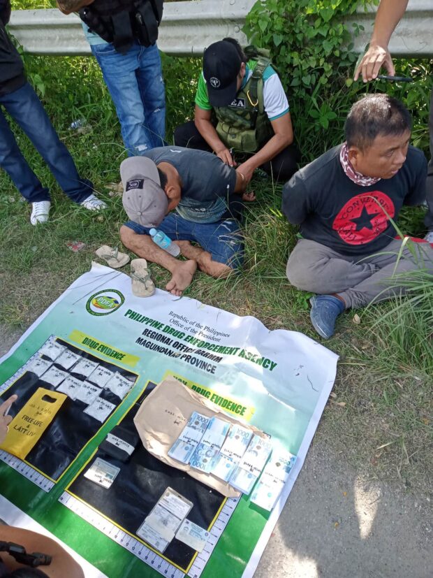 BUSTED. The two suspects sit while PDEA agents conduct an accounting of illegal stuff seized from them during a drug buy-bust operation in Cotabato City Thursday, May 4, 2023. (PDEA photo)