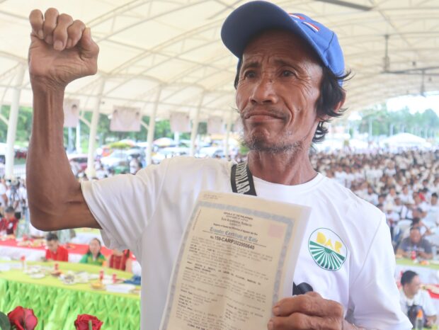 With fist up, an Agrarian Reform Beneficiary proudly shows his Certificate of Land Ownership Award, a dream come true after years of waiting, against the backdrop of thousand others who also get the same. Photo courtesy of DAR Agusan del Sur.