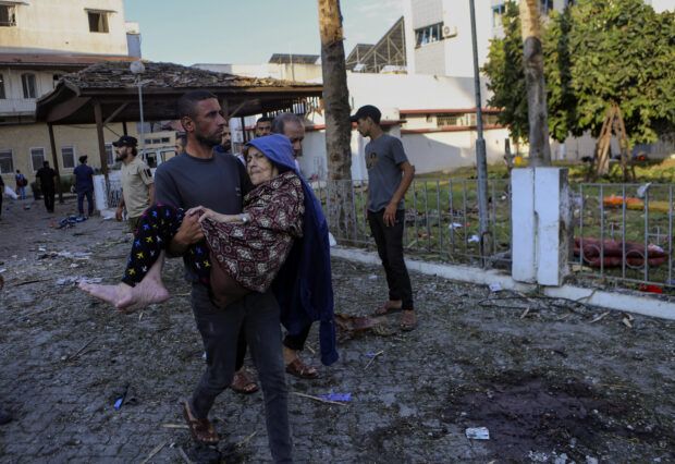 A Palestinian man carries an elderly woman past the site of a deadly explosion at al-Ahli hospital, in Gaza City, Wednesday, Oct. 18, 2023. 